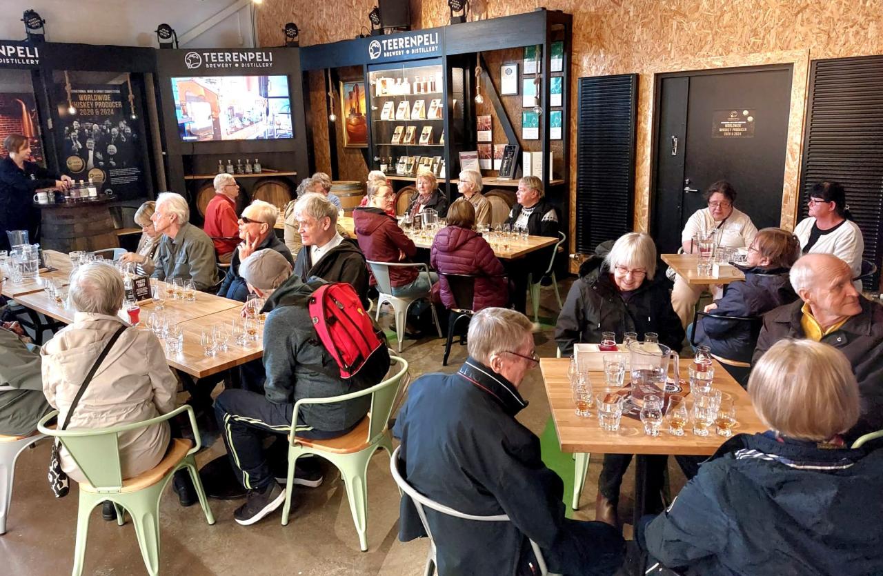 Kuva vieralijatilan ovelta, jossa näkyy etualalla yksi neljän hengen pöytä ja taustalla kaksi pitkää pöytää. Ihmisiä istuu pöytien ääressä juomalasit edessään ja osa katsoo vasemmassa kulmassa näkyvää esittelijää.